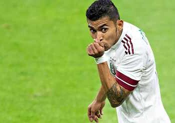 Jugador de fútbol en acción en el campo con uniforme blanco y tatuajes visibles.