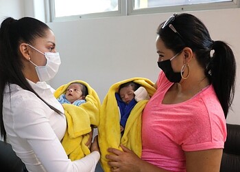 Nina recibiendo atención médica con madres con mascarillas, bebés envueltos en mantas amarillas.