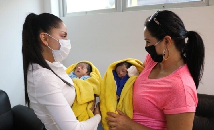 Nina recibiendo atención médica con madres con mascarillas, bebés envueltos en mantas amarillas.
