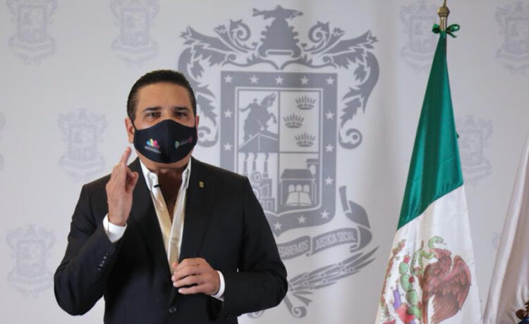 Muñeco con máscara y traje formal frente a escudo y bandera de México.