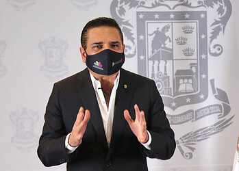 Prominente figura política mexicana con mascarilla y fondo oficial con bandera de México.