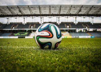 Balón de fútbol en campo de estadio vacío con gradas en el fondo.