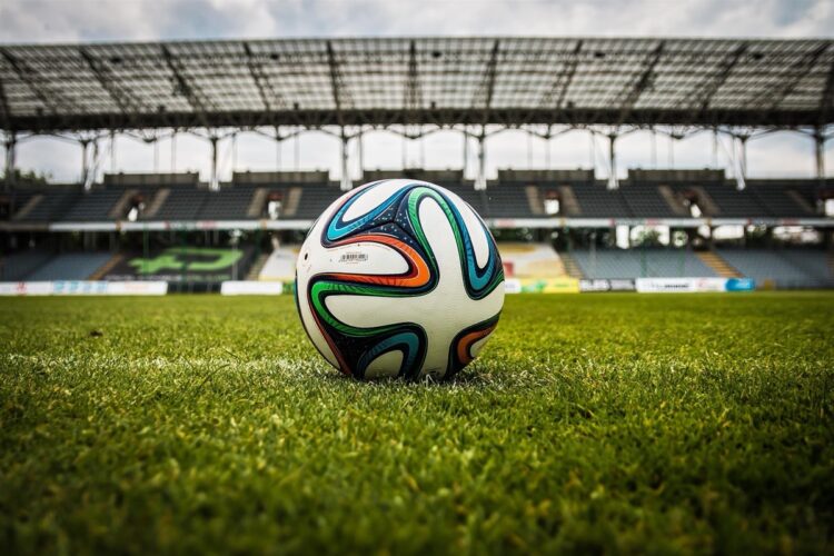 Balón de fútbol en campo de estadio vacío con gradas en el fondo.