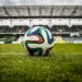 Balón de fútbol en campo de estadio vacío con gradas en el fondo.