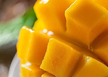 Fruta madura y jugosa en primer plano, con un fondo difuso de mangos frescos y hojas verdes.