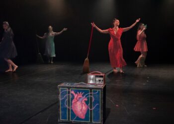 Bailarinas en escena con vestuarios coloridos y accesorios, realizando una presentación artística en un teatro oscuro.