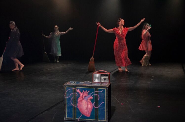 Bailarinas en escena con vestuarios coloridos y accesorios, realizando una presentación artística en un teatro oscuro.
