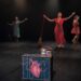 Bailarinas en escena con vestuarios coloridos y accesorios, realizando una presentación artística en un teatro oscuro.