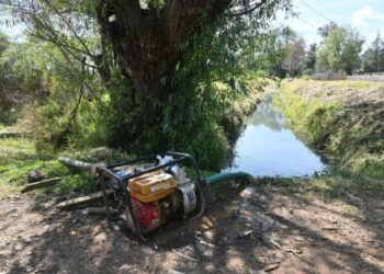 Bomba de agua robada junto a un canal con árboles y vegetación, evidencia de huachicol de agua en área rural.