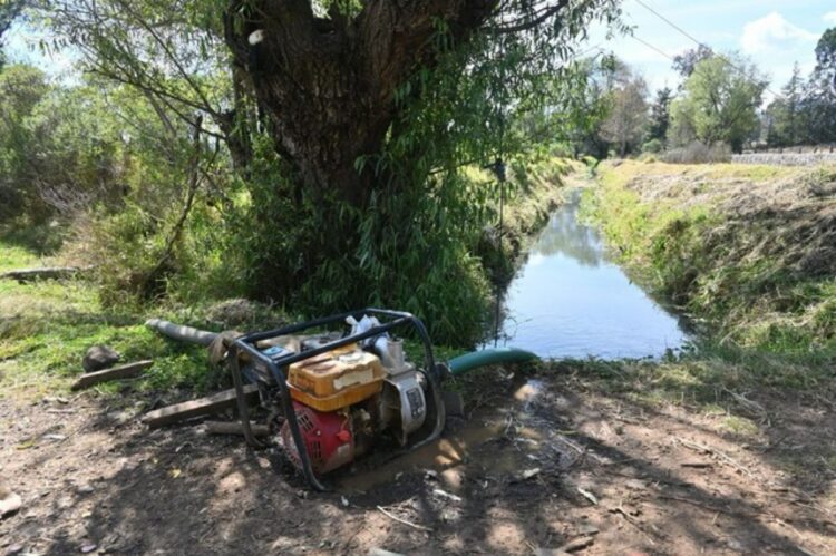 Bomba de agua robada junto a un canal con árboles y vegetación, evidencia de huachicol de agua en área rural.