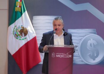 Auto en Michoacán, con bandera mexicana y fondo de monedas y símbolo de dinero, en una conferencia de prensa.