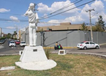 Estatua de una figura femenina en un parque urbano con coches y cables eléctricos en el fondo.