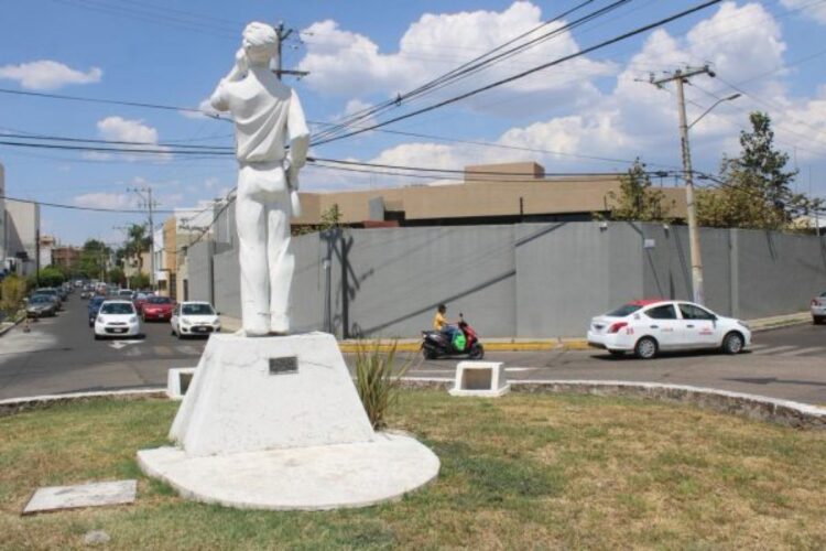 Estatua de una figura femenina en un parque urbano con coches y cables eléctricos en el fondo.