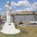 Estatua de una figura femenina en un parque urbano con coches y cables eléctricos en el fondo.