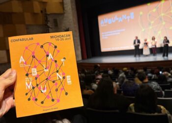 Cartel de Ambulante 2026 en mano, con el escenario y audiencia en el fondo en un evento cultural en Michoacán.