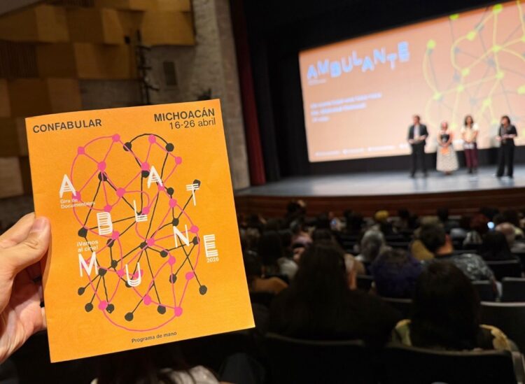 Cartel de Ambulante 2026 en mano, con el escenario y audiencia en el fondo en un evento cultural en Michoacán.