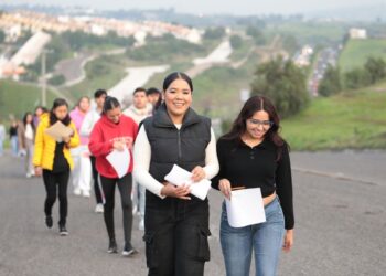 Estudiantes con documentos en mano caminando en un camino rural con paisaje de colinas y vegetación, en un día soleado.