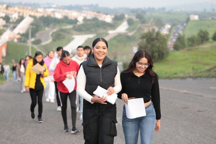 Estudiantes con documentos en mano caminando en un camino rural con paisaje de colinas y vegetación, en un día soleado.