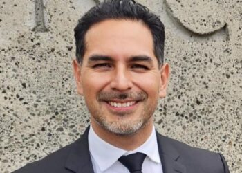 Hombre sonriente en traje formal con fondo de muro de piedra.