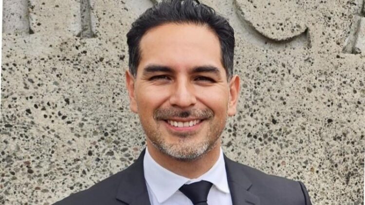 Hombre sonriente en traje formal con fondo de muro de piedra.