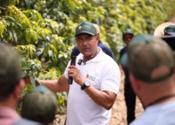 Hombre hablando con grupo de agricultores en un campo de cultivo, usando gorra y camiseta blanca, en un entorno agrícola.