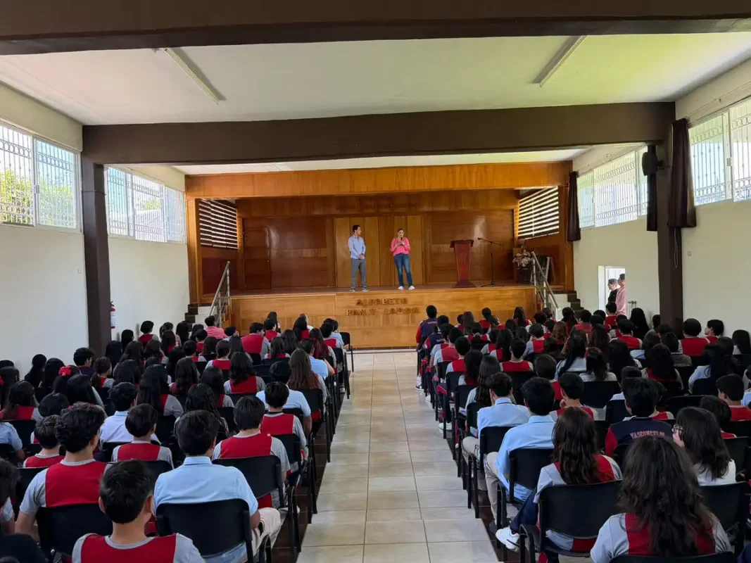 Estudiantes en evento escolar en auditorio, con dos presentadores en el escenario y audiencia atenta.