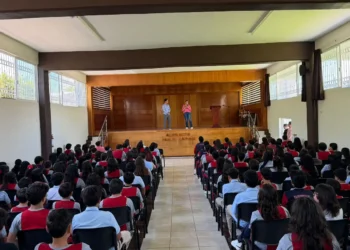 Estudiantes en evento escolar en auditorio, con dos presentadores en el escenario y audiencia atenta.