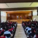 Estudiantes en evento escolar en auditorio, con dos presentadores en el escenario y audiencia atenta.