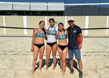 Jóvenes atletas femeninas en uniforme de voleibol en la arena junto a su entrenador en la playa.