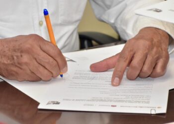 Persona firmando un documento importante con un bolígrafo en una oficina, con otra mano señalando el papel.