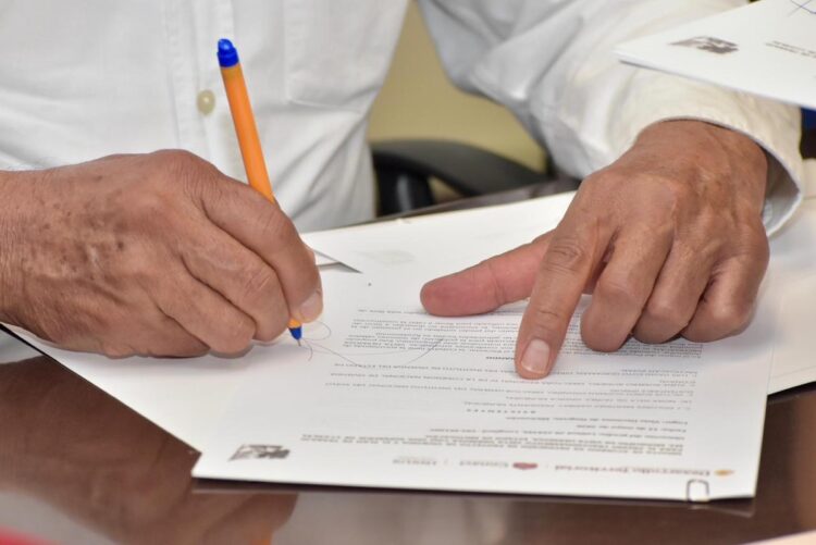 Persona firmando un documento importante con un bolígrafo en una oficina, con otra mano señalando el papel.