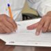 Persona firmando un documento importante con un bolígrafo en una oficina, con otra mano señalando el papel.