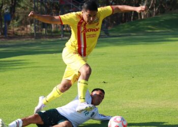 Jugador en uniforme amarillo saltando sobre oponente en campo de fútbol durante partido en exteriores.