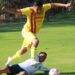 Jugador en uniforme amarillo saltando sobre oponente en campo de fútbol durante partido en exteriores.