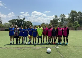 Jugadoras de fútbol femenino en entrenamiento en un campo al aire libre con árboles y cielo despejado de fondo.