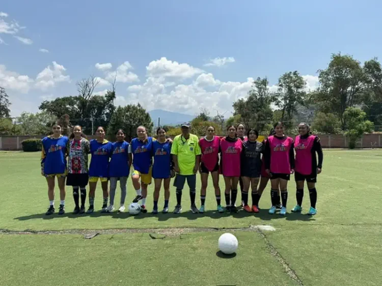 Jugadoras de fútbol femenino en entrenamiento en un campo al aire libre con árboles y cielo despejado de fondo.