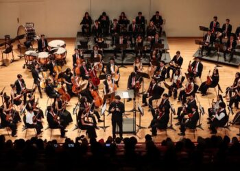 Orquesta sinfónica tocando en un escenario amplio con músicos en uniforme y director en el centro.