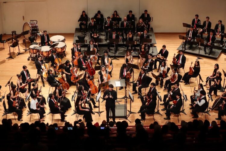 Orquesta sinfónica tocando en un escenario amplio con músicos en uniforme y director en el centro.