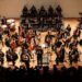 Orquesta sinfónica tocando en un escenario amplio con músicos en uniforme y director en el centro.