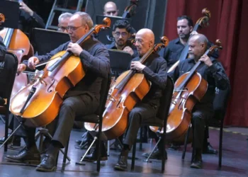Banda de músicos tocando violonchelos en un concierto en un escenario con cortinas rojas.