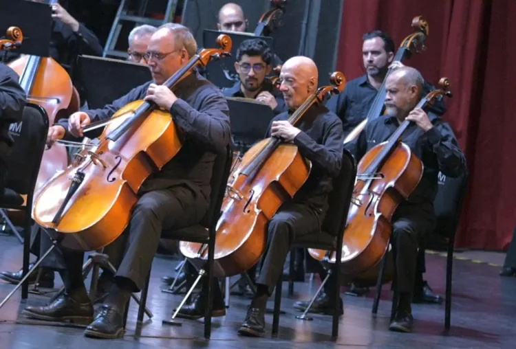 Banda de músicos tocando violonchelos en un concierto en un escenario con cortinas rojas.