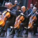 Banda de músicos tocando violonchelos en un concierto en un escenario con cortinas rojas.