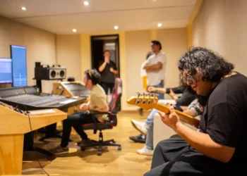 Músico tocando guitarra en estudio de grabación con equipo profesional y otros artistas en fondo.