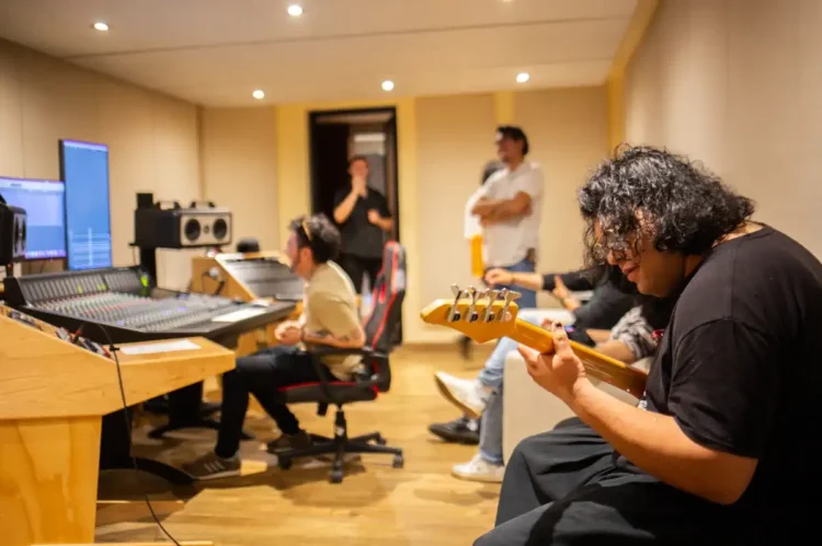 Músico tocando guitarra en estudio de grabación con equipo profesional y otros artistas en fondo.