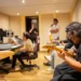 Músico tocando guitarra en estudio de grabación con equipo profesional y otros artistas en fondo.