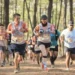 Participantes corriendo en una carrera de trail en un bosque, con árboles altos y luz solar filtrándose entre las hojas.