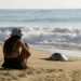 Mujer sentada en la playa observando una tortuga marina en la arena, con el mar y las olas de fondo, en un día soleado y tran.