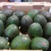 Aguacates verdes en caja de cartón en un mercado de Michoacán, con fondo difuso de otros productos y puestos.