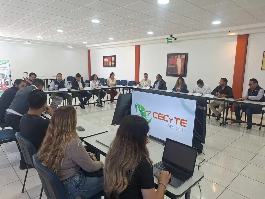 Grupo de personas en reunión en una sala moderna con pantallas y laptops, discutiendo temas educativos en Michoacán.