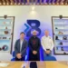 Tres hombres en una sala con estanterías llenas de objetos decorativos y un logo grande en la pared.